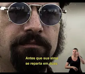 Close do rosto de um homem branco de pele coberta por sardas, longas suíças, bigode e cavanhaque castanhos. Usa óculos escuros de aros redondos. Na base da tela, em caracteres amarelos, a legenda: “Antes que sua alma se reparta em duas.” No canto inferior direito, a intérprete Ângela Russo sinaliza.