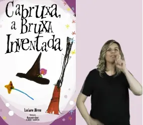 Fundo lilás claro. À esquerda está a capa do livro “Cabruxa, a Bruxa Inventada”, de Luciana Abreu. A capa do livro é branca, com bordas em lilás. O título está escrito no alto, com letras pretas e tremidas, de diferentes tamanhos. No centro estão desenhadas três figuras: um chapéu de bruxa, preto e pontudo, enfeitado por uma flor lilás; uma varinha de condão com uma estrela amarela na ponta; e uma vassoura de palha. As margens são cheias de estrelinhas coloridas e círculos brancos e lilases que parecem flutuar.À direita, a intérprete  ngela Russo faz o sinal de “bruxa”, com o indicador em gancho ao lado do nariz.