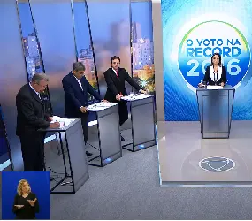 Cinco candidatos à prefeitura de Porto Alegre em estúdio de TV. Estão atrás de púlpitos cinza com laterais vazadas, posicionados em semicírculo, com três candidatos à esquerda e dois à direita. No centro, a mediadora, também atrás de um púlpito. Atrás dela, um painel azul com a identidade gráfica do programa: O Voto na Record – 2016. No canto inferior esquerdo, a intérprete Ângela Russo na janela de Libras