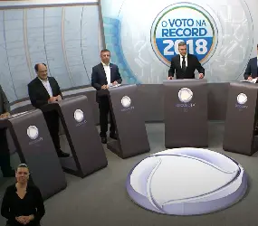 Seis candidatos ao governo do estado do RS em estúdio de TV. Estão atrás de púlpitos cinza escuro posicionados em semicírculo, com três candidatos à esquerda e três à direita. Entre eles, também atrás de um púlpito, está o mediador. Atrás dele, um painel com a identidade gráfica do programa: O Voto na Record – 2018. Os candidatos vestem trajes esporte e o mediador usa terno e gravata. No canto inferior esquerdo, a intérprete  ngela Russo na janela de Libras. No canto inferior direito, o símbolo da emissora e a identificação ao vivo